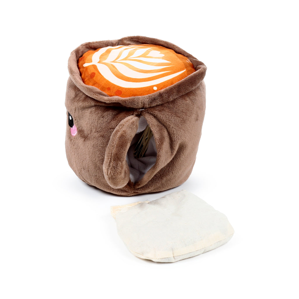 Plush brown heat pack, shaped like a coffee cup character, featuring an embroidered eye and pink cheek. Its open top reveals orange 'latte' art, and a separate cream fabric heat pack insert lies next to it on a white background.