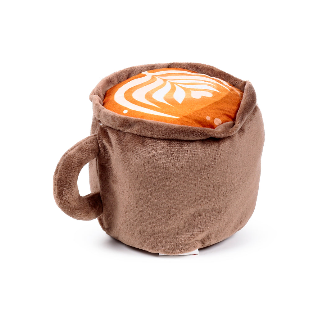 Angled view of a brown plush microwavable heat pack shaped like a cappuccino mug, featuring a white latte art pattern on its foamy top surface.