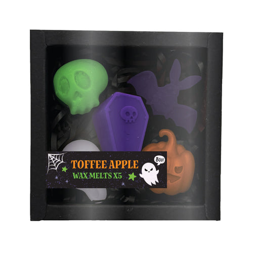 Front view of five Halloween-themed wax melts in a black box with a clear window and black shredded filler. The melts include a green skull, a purple coffin, a purple bat, an orange pumpkin, and a white dome shape. A label on the box advertises 'Toffee Apple Wax Melts x5' and features a white ghost and a spiderweb design.