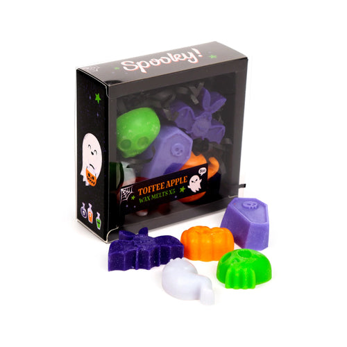 Angled view of a 'Spooky!' themed toffee apple wax melt set, featuring a black display box with a clear front. Visible inside the box are green skull, purple bat, and purple coffin wax melts amidst shredded paper. Five individual spooky-shaped wax melts are arranged in front: a purple bat, orange pumpkin, white ghost, green spider, and purple coffin, all on a white background.