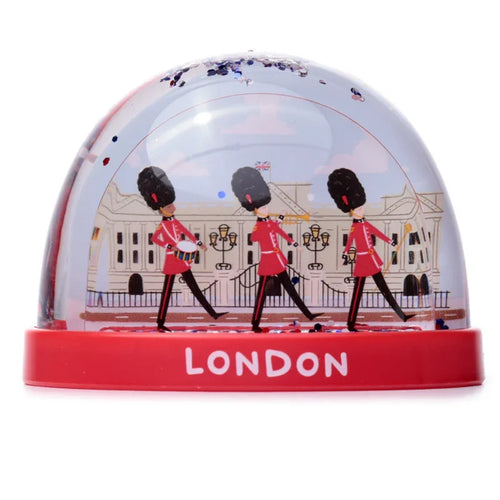 Front view of a London glitter snow globe, featuring three marching Queen's Guards in red uniforms and bearskin hats, with a palace-like building behind them and 'LONDON' text on the red base.