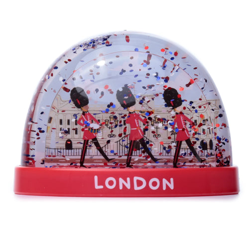 Front view of a dome-shaped snow globe, featuring three British Queen's Guards marching against an illustrated building. Red, blue, and white glitter fills the clear dome, and the red base displays 'LONDON' in white capital letters.
