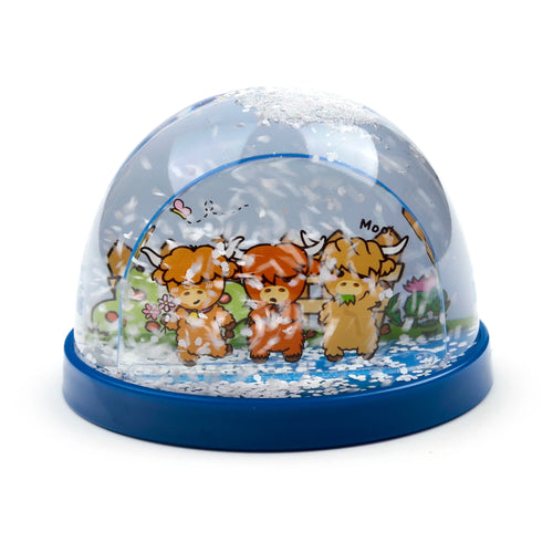 Angled view of a glitter snow globe, featuring three cartoon Highland cows; one holds flowers, another eats grass, set against a green and blue landscape with a 'Moo!' bubble. The globe contains white glitter flakes and rests on a dark blue base.
