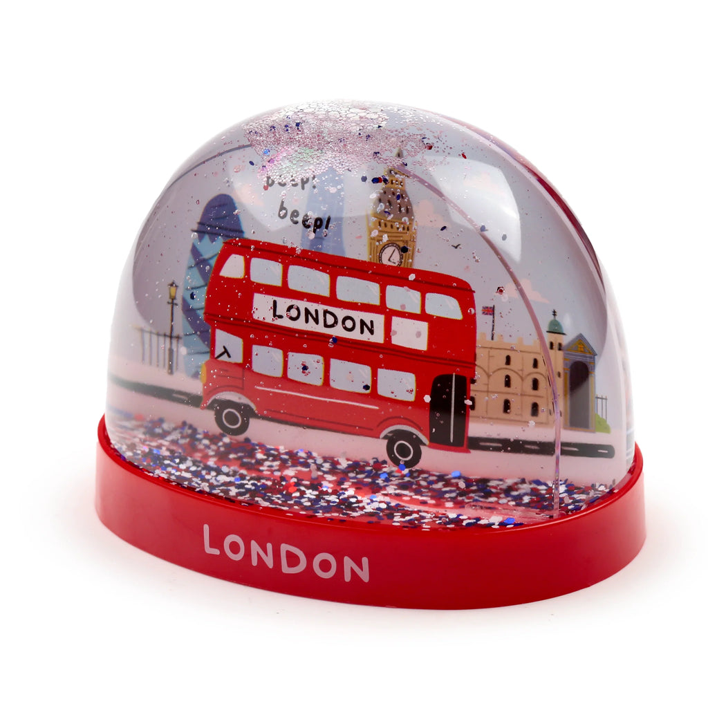 Angled view of a London bus snow globe, featuring a red double-decker bus, Big Ben, and other London landmarks. The snow globe contains red, white, and blue glitter, and sits on a red base with 'LONDON' written on it.