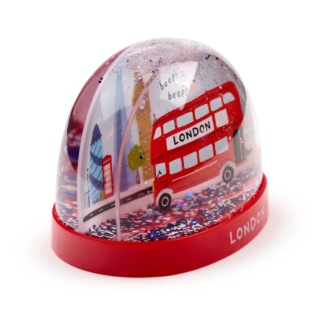 Angled view of a glitter snow globe, depicting a red London double-decker bus, Big Ben, and The Gherkin, with red, white, and blue glitter. It rests on a red base inscribed with 'LONDON'.
