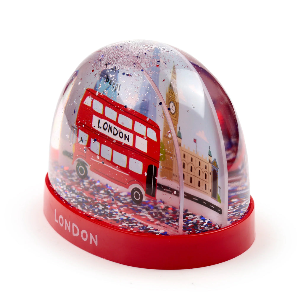 Angled view of a snow globe with a red base. Inside the clear dome, a miniature red double-decker London bus is shown alongside an illustration of Big Ben. Multi-coloured glitter is suspended within the globe, and 'LONDON' is visible on the red base.
