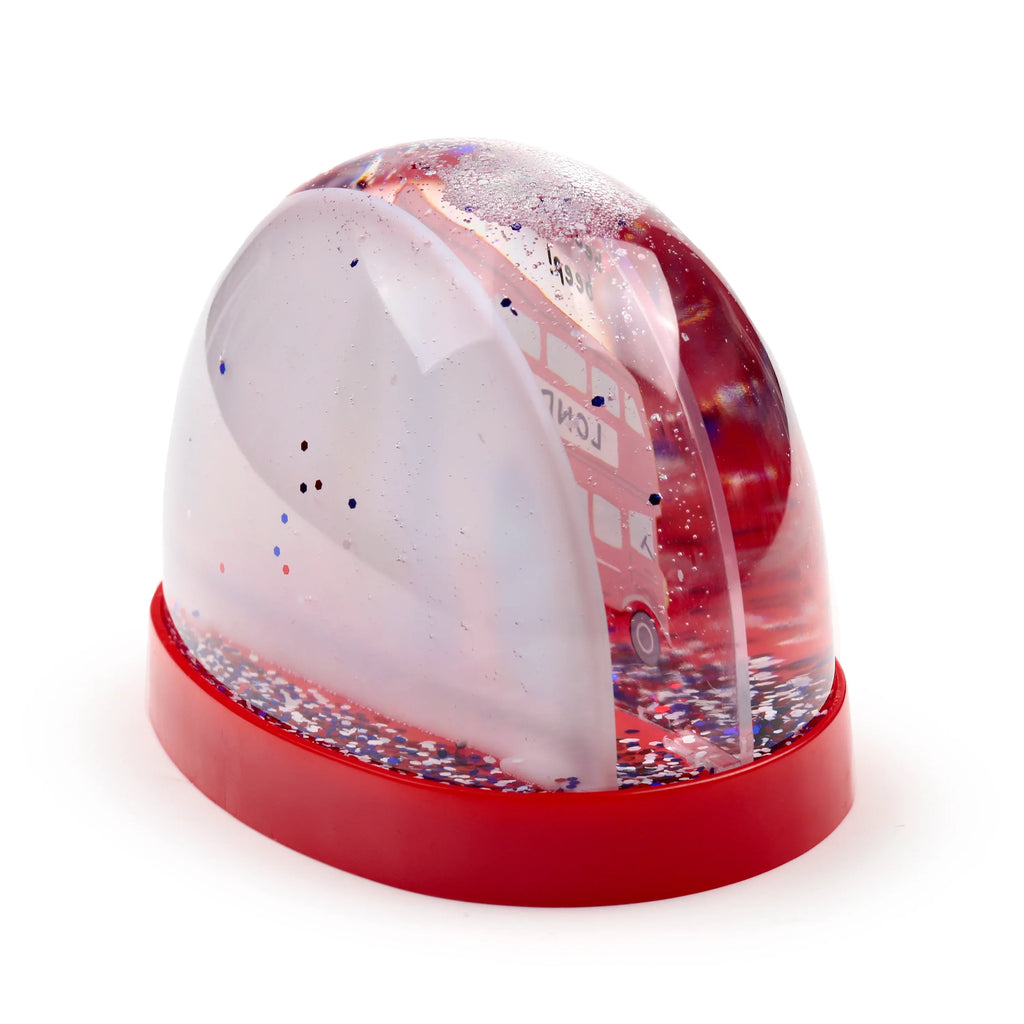 Angled view of an oval glitter snow globe with a red London bus inside. It has a clear dome containing red, white, and blue glitter, set on a red oval base.