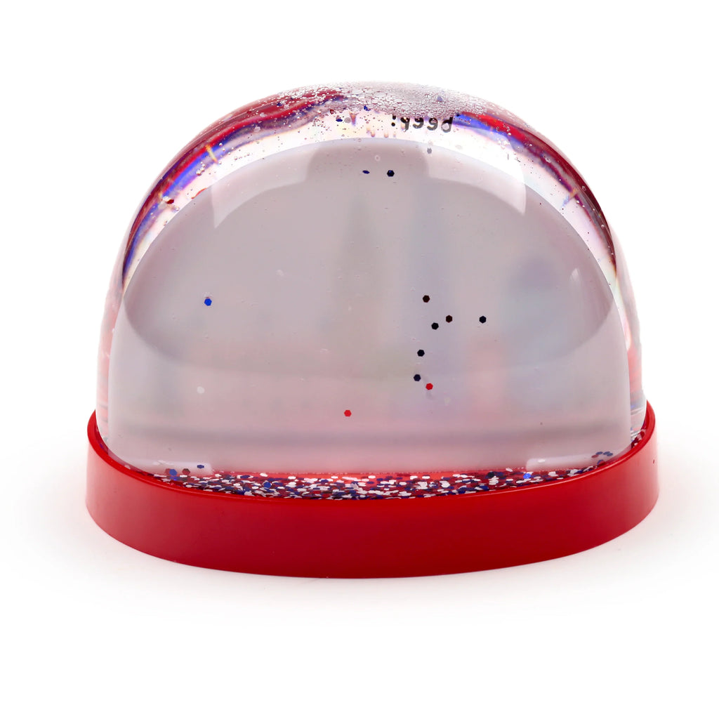 Angled view of a snow globe with a clear dome and a red base, containing red, white, and blue hexagonal glitter suspended in the liquid and settled on the base.
