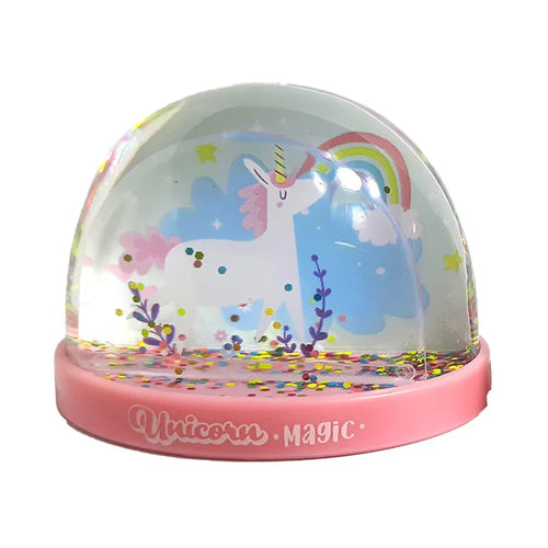 Front view of a snow globe featuring a white unicorn with pink mane and tail, set against a pastel blue background with a rainbow and clouds. The dome contains multicoloured glitter, and the pink base displays 'Unicorn Magic'.