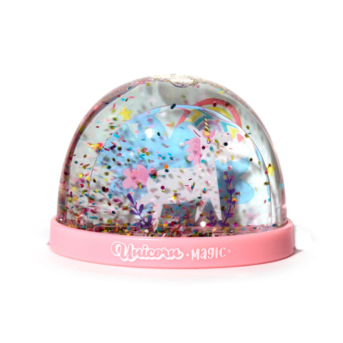 Front view of a Unicorn Magic snow globe, featuring a white unicorn with pink mane and tail, green grass, pink flowers, purple foliage, a rainbow, and white clouds inside a clear dome filled with multicoloured glitter, set on a pink base with Unicorn Magic text.