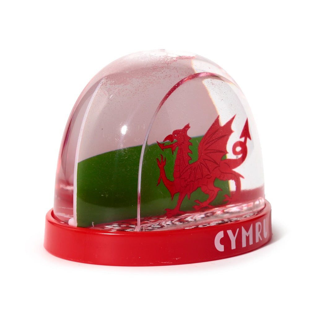 Angled front view of a collectable Welsh dragon snow globe, featuring a red dragon on a green background within the clear dome, with visible snow-like particles and 'CYMRU' text on the red base.
