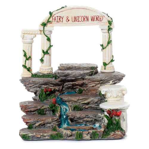 Front view of a Fairy & Unicorn World figures display stand, depicting a rock-effect tiered landscape with a blue waterfall, white columns, and an archway bearing 'Fairy & Unicorn World' text, adorned with green ivy, grass, and red flowers.