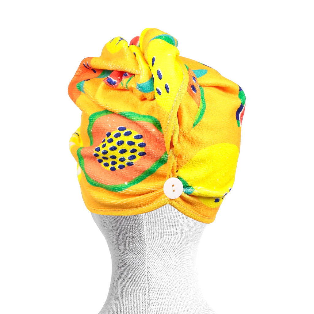 Angled view of a yellow hair wrap towel with a vibrant summer fruits pattern, including papaya and watermelon, fastened by a white button, on a white mannequin.
