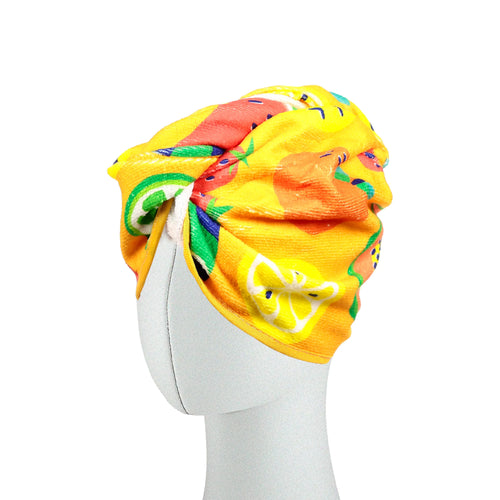 Angled view of a yellow, turban-style hair wrap towel with a colourful summer fruit pattern, modelled on a mannequin head.