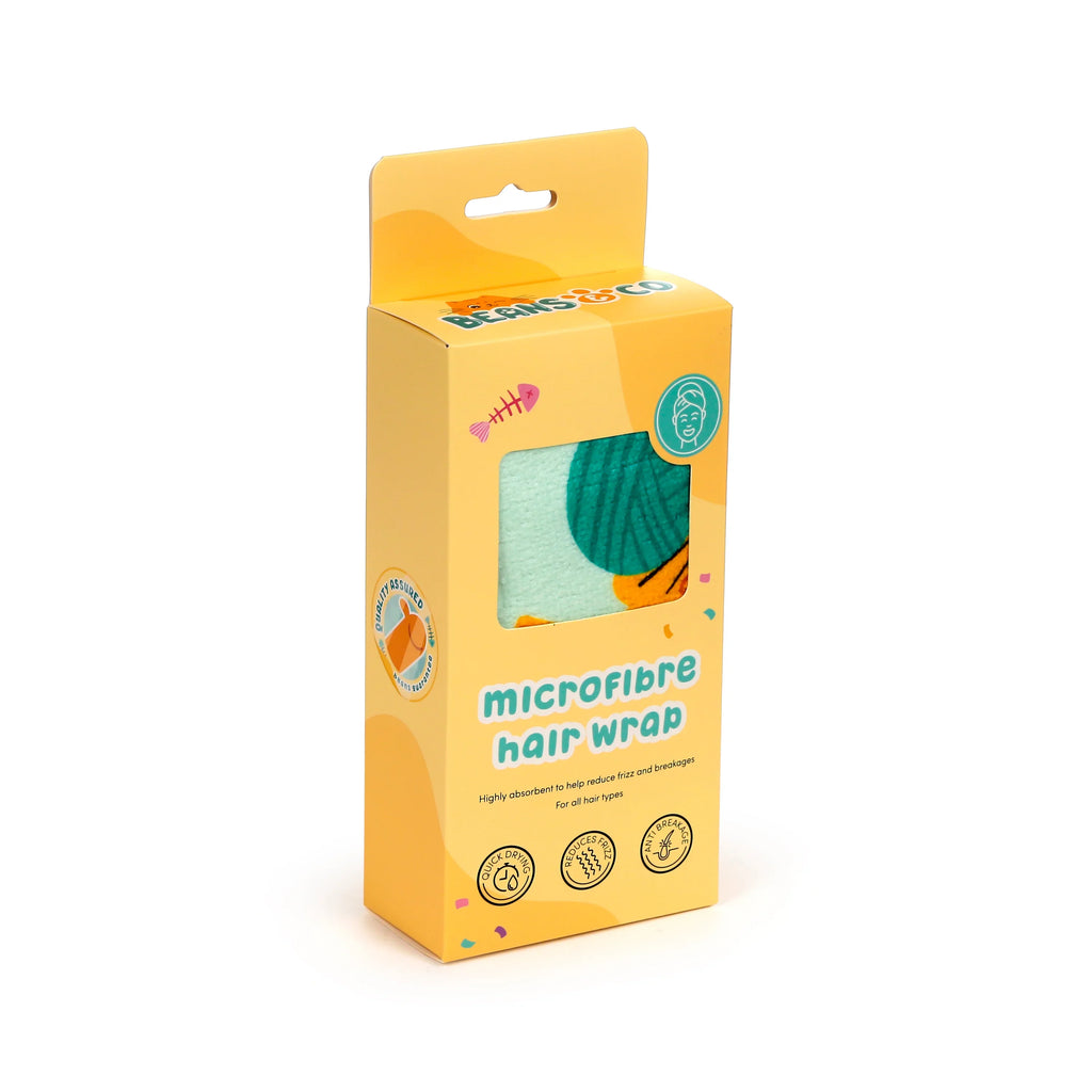 Angled view of a bright yellow microfibre hair wrap box with a hang tab, featuring playful cat and fish bone graphics, and text highlighting its quick-drying, frizz-reducing, anti-breakage benefits. A window reveals a partial view of the green and orange cat-themed towel inside.