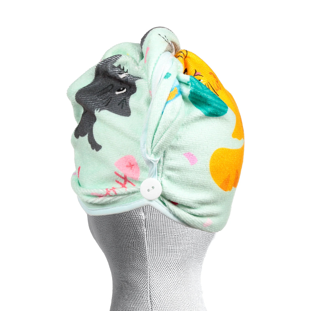 Angled view of a mint green hair wrap towel with a playful cat and fishbone pattern, featuring black and orange cats. A white button on the front secures the towel, which is displayed on a grey mannequin head.