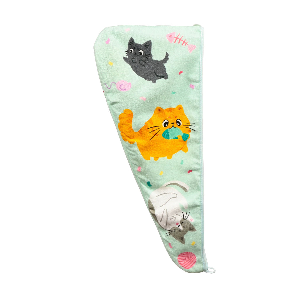 Overhead view of a light green hair wrap towel displaying a playful cartoon cat pattern. It features a black cat, an orange cat holding a blue fish, a grey and white cat with a pink yarn ball, colourful confetti, and a pink fish skeleton.