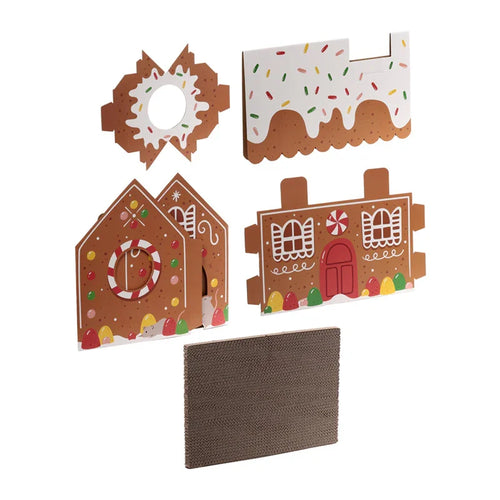 Overhead view of unassembled brown cardboard cat den playhouse components, featuring gingerbread house panels decorated with white icing and colourful candy details, alongside a corrugated scratching pad.