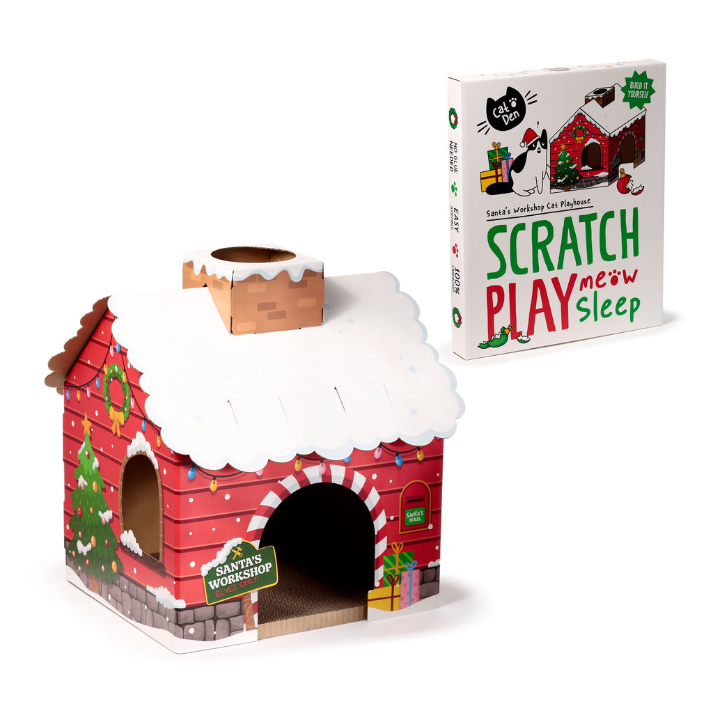 Angled view of a festive red cardboard cat den playhouse, styled as a Christmas grotto, with a white snowy roof, a chimney, an arched entrance, a Christmas tree, wreath, and colourful string lights. Its white packaging box, displaying 'Scratch Play Sleep' and a cartoon cat, is visible in the background.
