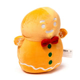Angled front view of an orange plush gingerbread man, featuring white icing details, a red bow tie, and three green buttons.