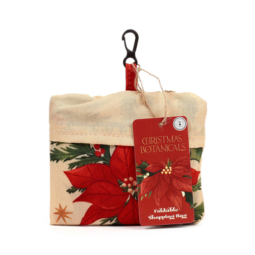 A compact, folded shopping tote bag depicting a Christmas botanical pattern with red poinsettias, green leaves, and berries on a cream background. A black karabiner clip is attached at the top via a red fabric loop. A red product tag hangs from twine, displaying 'Christmas Botanicals Foldable Shopping Bag' and a 'Recycled Plastic' logo.