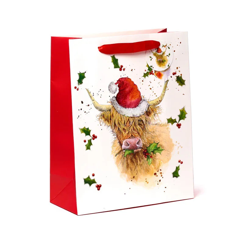 Angled view of a large white Christmas gift bag with red sides and red ribbon handles. The front features an illustration of a Highland Coo wearing a red Santa hat and holding green holly, surrounded by scattered holly leaves and red berries.