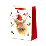 Front view of a large white gift bag with a red side panel and handles, featuring a Christmas illustration of a Highland Coo wearing a Santa hat, munching holly, with a robin and scattered holly leaves.