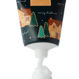 Close-up of a moisturising hand cream tube, showing white cream being dispensed from its nozzle. The dark tube is decorated with festive gingerbread houses and green Christmas trees, featuring merry christmas text and a visible 75ml volume indicator.