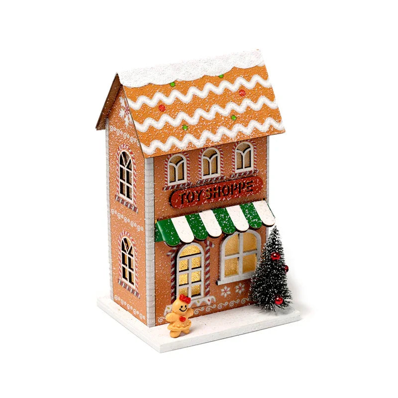Front view of an LED Christmas gingerbread toy shoppe decoration, with brown walls, white icing details, illuminated windows, a green and white striped awning, a 'Toy Shoppe' sign, a small Christmas tree with red baubles, and a gingerbread person figure on a snow-effect base.