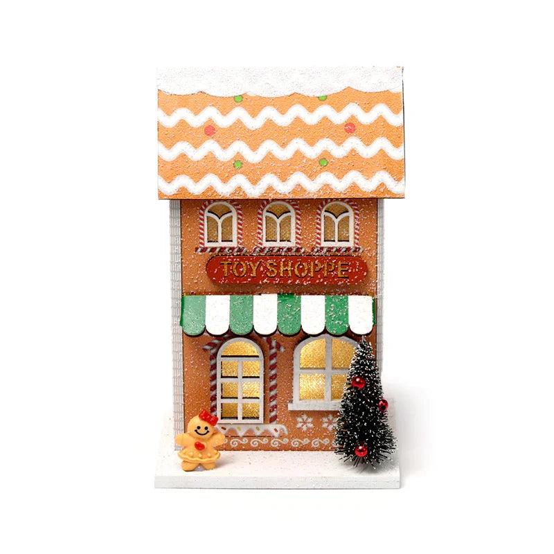Front view of an illuminated LED Christmas decoration, a gingerbread-effect toy shoppe. It features a snow-dusted roof, a red 'Toy Shoppe' sign, a green and white striped awning, a miniature Christmas tree with red baubles, and a gingerbread person figure.