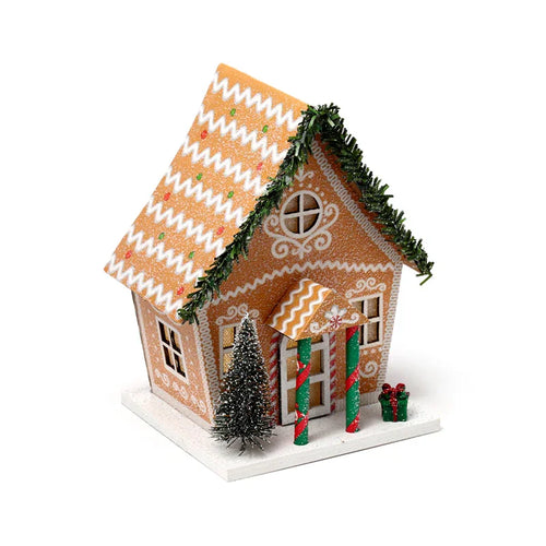 Angled view of an LED Christmas gingerbread house decoration. The orange house features glowing windows, white icing details, a roof with white zigzag patterns and coloured flecks, green tinsel garland, a miniature Christmas tree, and a small green gift box, all on a white snow-effect base.