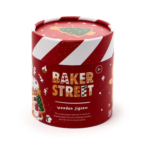 Front, angled view of a red cylindrical wooden jigsaw puzzle tube. The Christmas-themed container features white candy cane stripes, gingerbread illustrations, 'BAKER STREET' text, and a green Christmas tree on its lid.