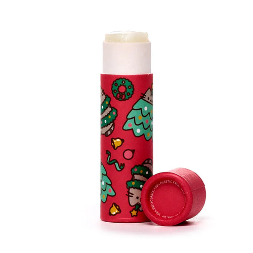 Angled view of a paper stick lip balm, showing a red tube decorated with Christmas-themed illustrations of Pusheen the Cat characters, green Christmas trees, wreaths, and gold bells. A pale yellow lip balm stick is extended from the top of the tube. The matching red cap lies detached next to the tube, with 100% RECYCLABLE. 100% PLASTIC FREE. text visible on its base.