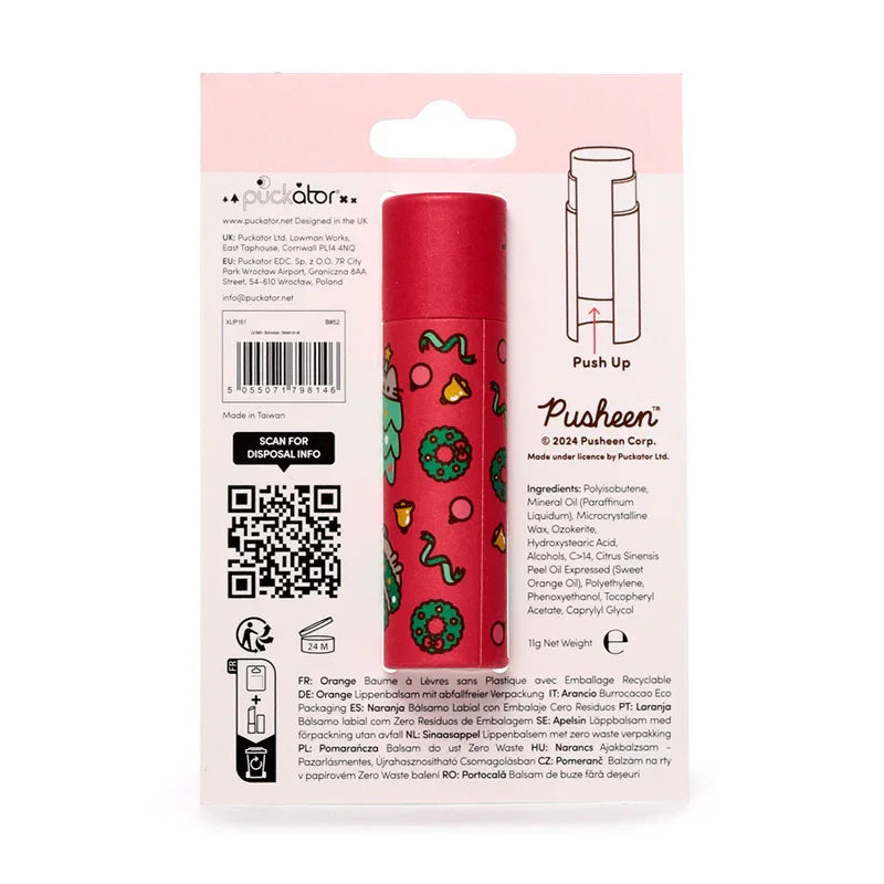 Back of a Christmas Pusheen the Cat paper stick lip balm on its white retail packaging. The red lip balm stick is decorated with green festive wreaths, bells, baubles, and a light blue Pusheen cat wearing a Santa hat. Visible on the packaging are 'Push Up' instructions, an ingredients list, and multilingual descriptions for an orange-scented, zero-waste lip balm.