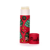 Upright paper stick lip balm with translucent pale yellow balm extended, its red cap removed and placed alongside. The red tube is decorated with a festive pattern of green Christmas trees, wreaths, gold bells, ribbons, and Pusheen the cat wearing a green Christmas tree hat. The word 'Pusheen' is visible on the cap.