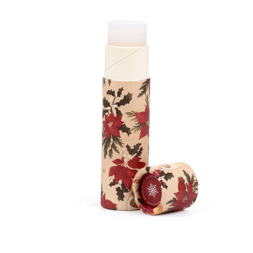 Angled view of a paper stick lip balm with Christmas botanicals design, showing the white lip balm exposed from the upright tube. The natural kraft paper tube and detached cap feature red poinsettias, green holly, and pine needles; the cap also has a red label with a white snowflake.