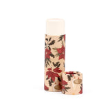Angled view of a paper stick lip balm. The upright main tube and its separate cap feature a Christmas botanicals pattern with red poinsettias, green holly leaves, and berries on a light beige background. The creamy lip balm is visible at the tube's opening.