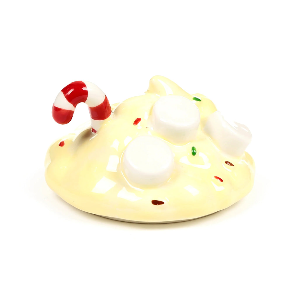 Angled view of a creamy yellow ceramic mug lid, decorated with white marshmallow shapes, colourful sprinkles, and a red and white candy cane.