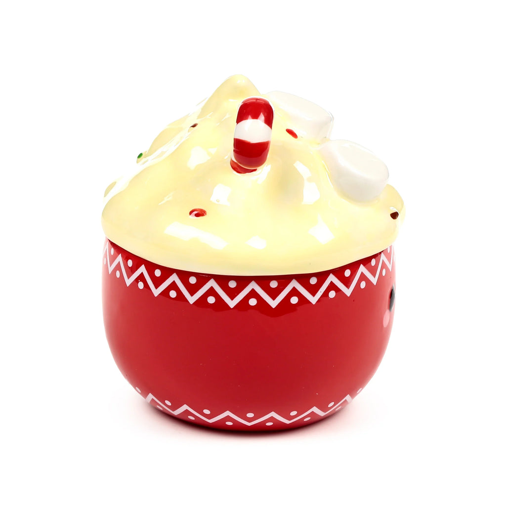 Angled view of a red ceramic lidded mug designed to resemble hot chocolate. The mug base features a white zig-zag pattern with dots around the rim and a character face design on one side. The pale yellow lid looks like whipped cream, topped with a red and white candy cane, white marshmallow shapes, and red sprinkles.