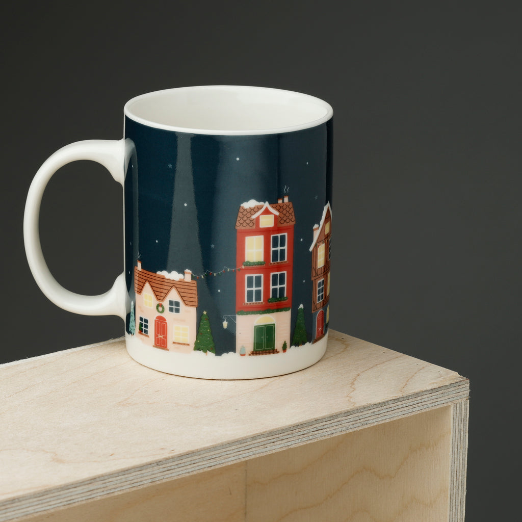 Angled view of a Christmas Village porcelain mug, with a dark blue exterior featuring an illustration of snow-covered houses, fir trees, and string lights, showing the white handle.