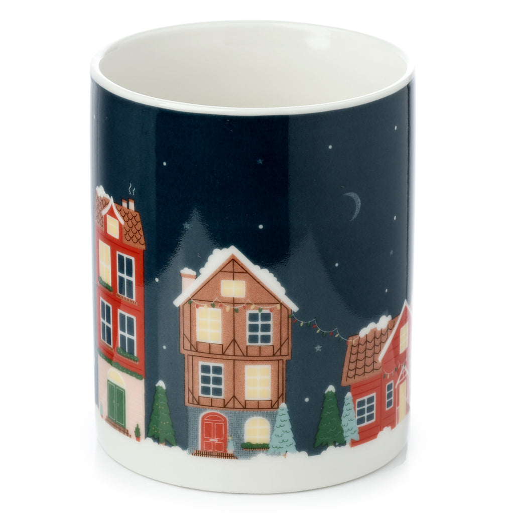Angled view of a porcelain mug with a white interior rim. Its dark blue exterior features a festive Christmas village scene, depicting colourful houses with illuminated windows, snow-covered ground, Christmas trees, a crescent moon, and stars in the night sky.