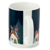 Angled view of a white porcelain Christmas village mug, featuring a dark blue winter scene with houses and a windmill covered in snow, displaying its white handle.