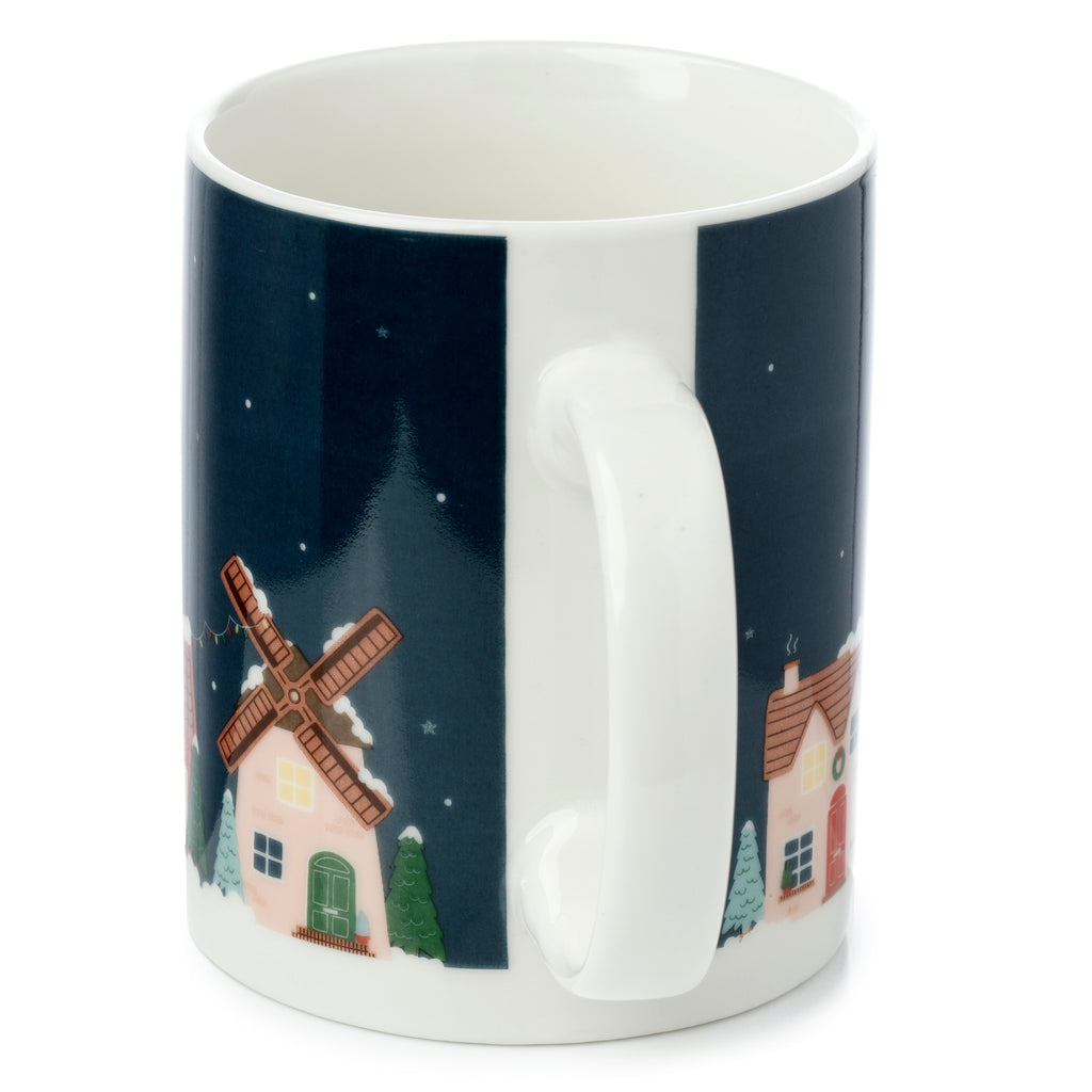 Angled view of a white porcelain Christmas village mug, featuring a dark blue winter scene with houses and a windmill covered in snow, displaying its white handle.