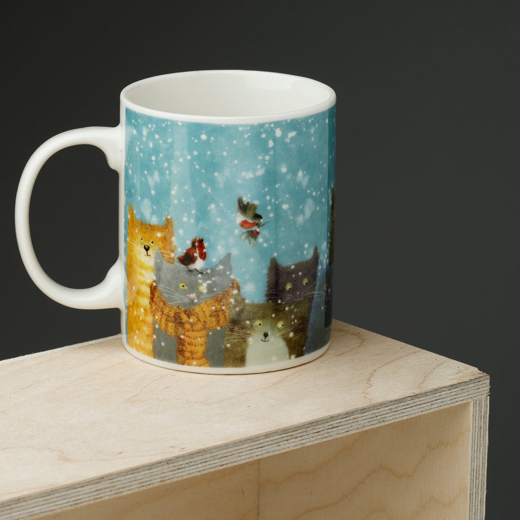 Angled view of a white porcelain mug with a Christmas illustration of several cats in a snowy light blue scene, featuring a robin and holly.