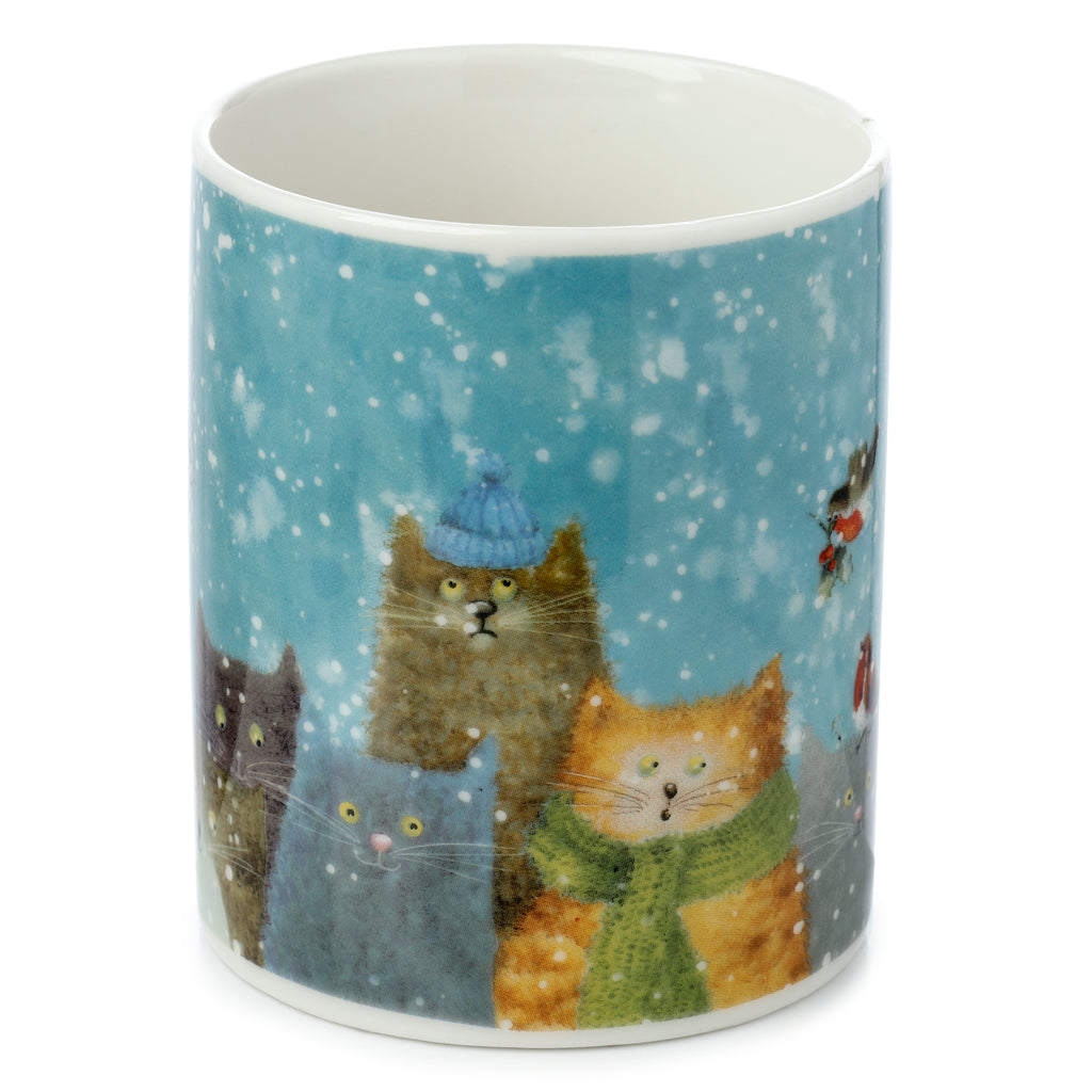 Angled view of a porcelain mug depicting a blue winter scene with white snow, featuring a brown cat in a blue hat, an orange cat in a green scarf, a grey cat, a black cat, and a small red-breasted robin.