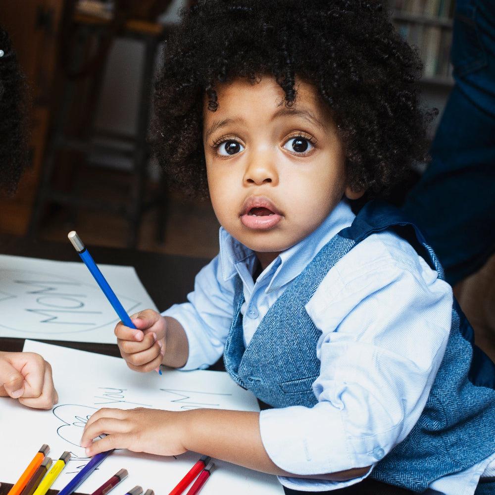 Child holding a blue pencil, sitting at a table with coloring books and crayons.