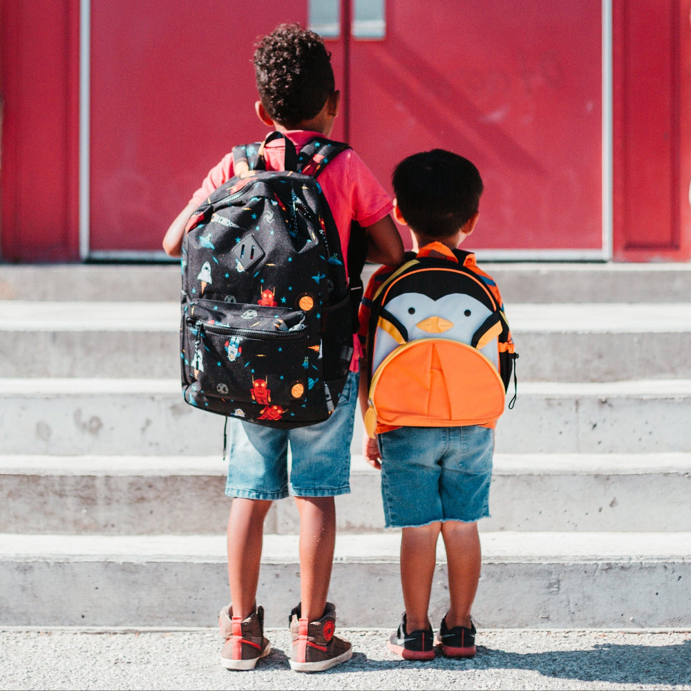 Two children with backpacks standing on steps in front of a red building.