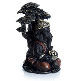 Black and gold Ganesh figurine sitting under a tree, showing an intricate gold crown, reddish-brown robes, and a bowl of offerings, from an angled view.