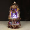 Front view of a backflow incense burner, featuring a skull with purple LED glowing eyes, illuminated crystals inside a glittery cave, and descending incense smoke over a textured, skeletal base.