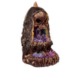 Angled view of a skull backflow incense burner, featuring a dark, textured rock formation with an exposed skull at the top. Inside, clear crystals are surrounded by a vibrant, glittery purple, blue, and yellow cave interior. A brown incense cone holder sits on top.